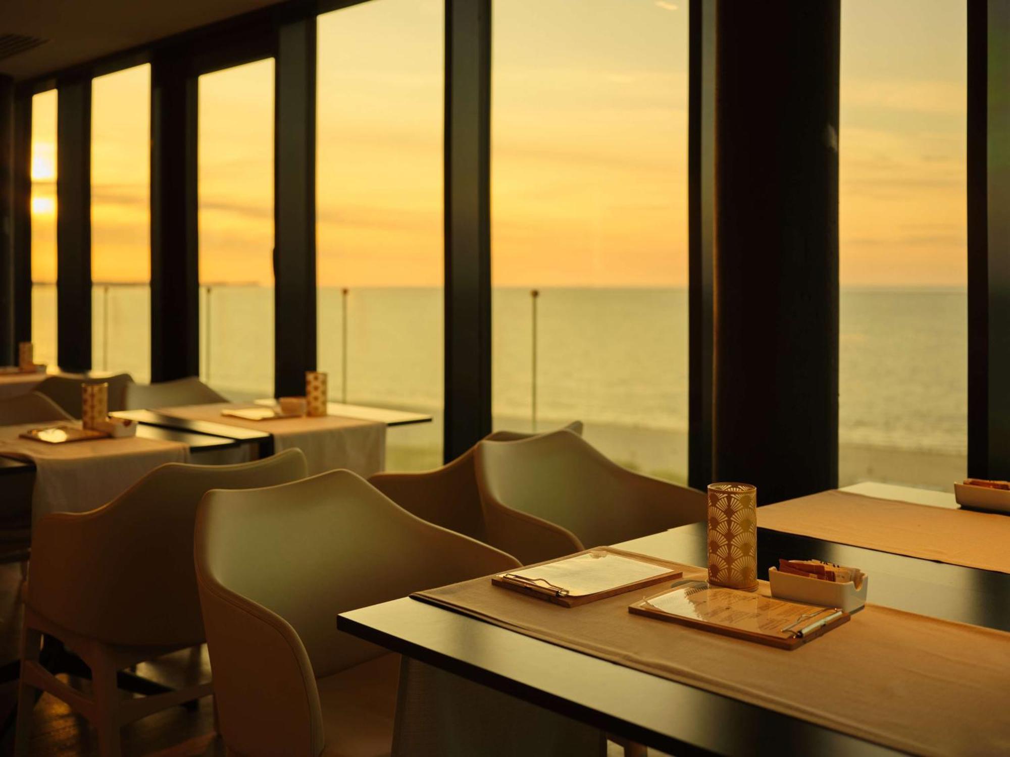 Radisson Blu Resort Świnoujście Екстериор снимка The photo shows a restaurant interior with a beautiful view of the ocean during sunset. The large windows allow natural light to flood the space, highlighting the warm colors of the sunset in the sky. There are several tables, each set with placemats