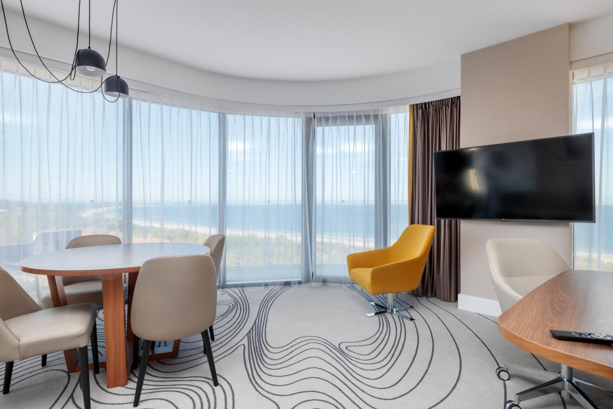 Radisson Blu Resort Świnoujście Екстериор снимка The image shows a modern hotel room or suite with a bright, airy atmosphere. There are large windows that allow natural light to fill the space, providing a view of the ocean. The decor includes a round dining table with several chairs, and a stylish