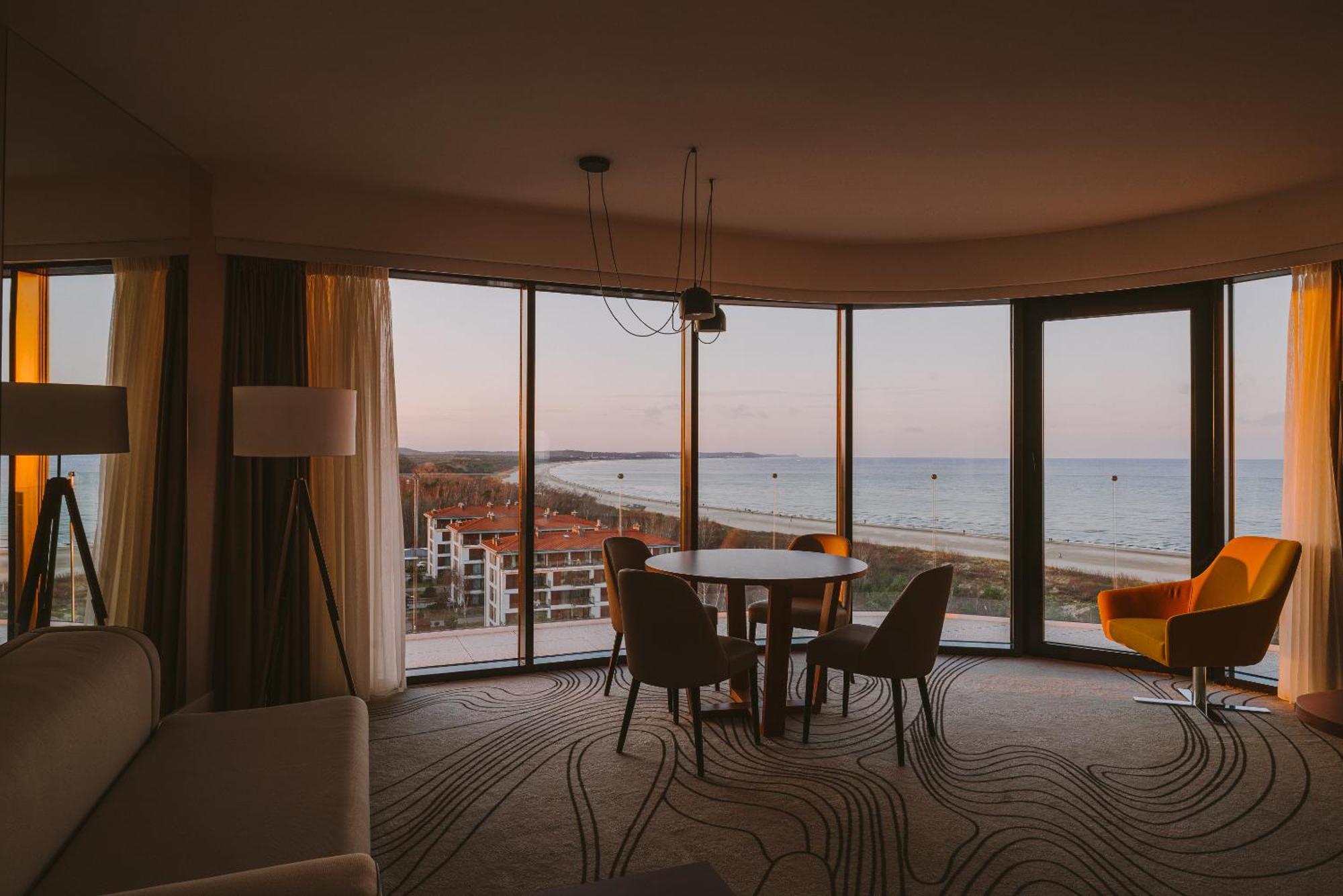 Radisson Blu Resort Świnoujście Екстериор снимка The photo shows a modern and stylish interior of a room featuring large windows that provide a view of the beach and ocean. The room is furnished with a round table and chairs, along with a cozy sofa and a bright yellow armchair. The decor has a cont