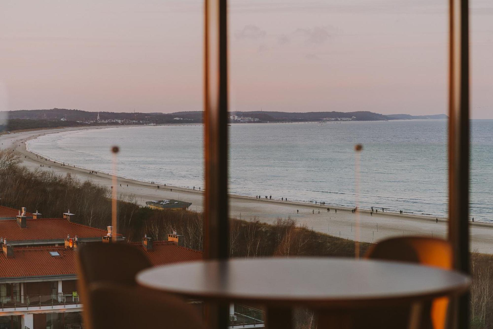Radisson Blu Resort Świnoujście Екстериор снимка The photo shows a picturesque view of a beach scene from indoors. In the foreground, there is a round table with some chairs around it, suggesting a cozy interior space. Through a large window, you can see the beach with gentle waves hitting the shor