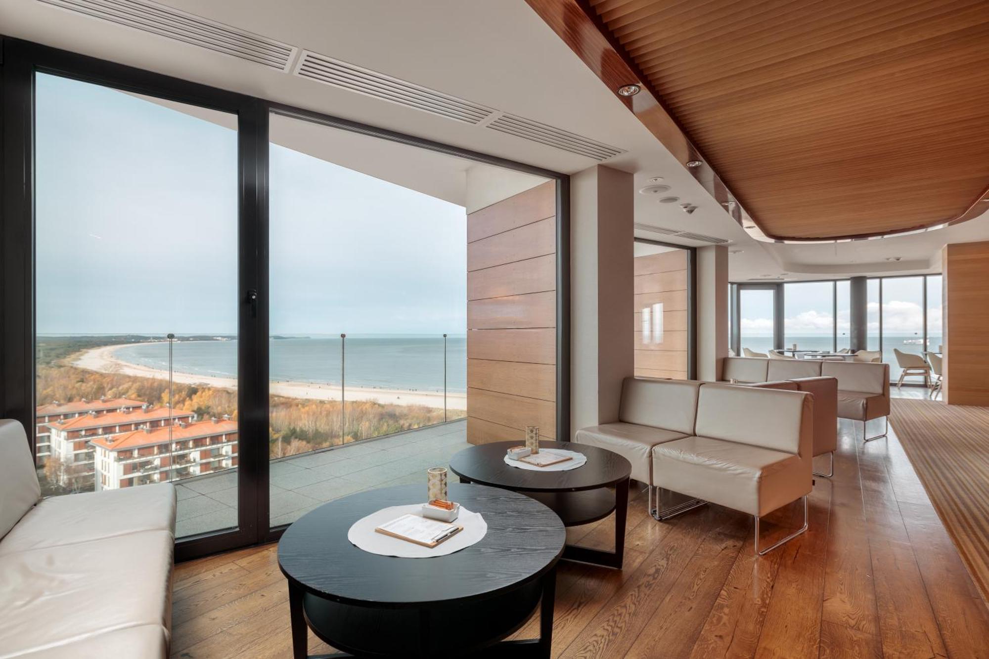 Radisson Blu Resort Świnoujście Екстериор снимка The photo shows a modern, elegantly designed interior space featuring large windows that offer a panoramic view of the ocean and coastline. The flooring is wooden, providing a warm atmosphere. There are two round black tables in the foreground with m