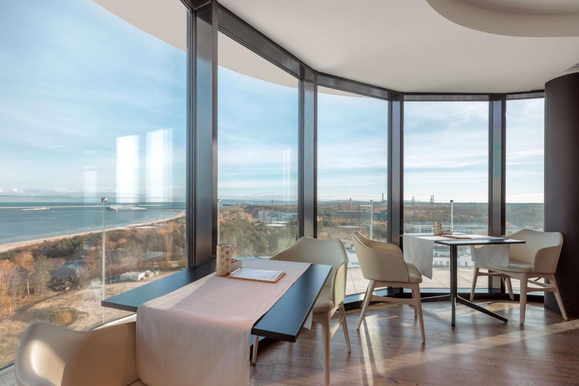 Radisson Blu Resort Świnoujście Екстериор снимка The photo shows a modern dining area with large windows that offer a panoramic view of the landscape outside, including a body of water and possibly a shoreline. The design features sleek furniture, including a table with chairs arranged around it, a