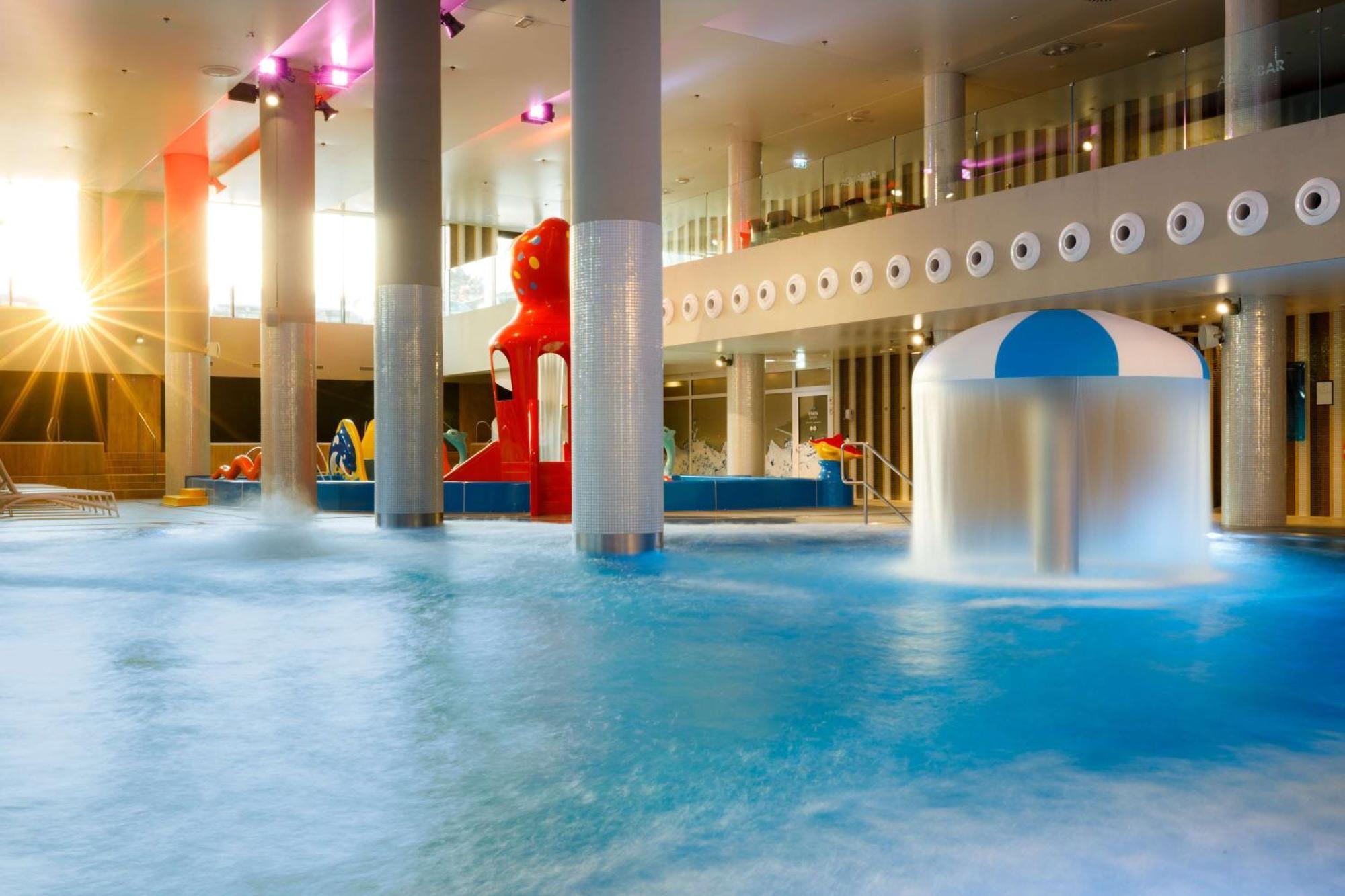 Radisson Blu Resort Świnoujście Екстериор снимка The photo shows an indoor water park or pool area. There is a large body of water with playful features, including a water slide shaped like a red bear and a smaller structure with a blue and white canopy. The lighting is bright, suggesting a sunny a