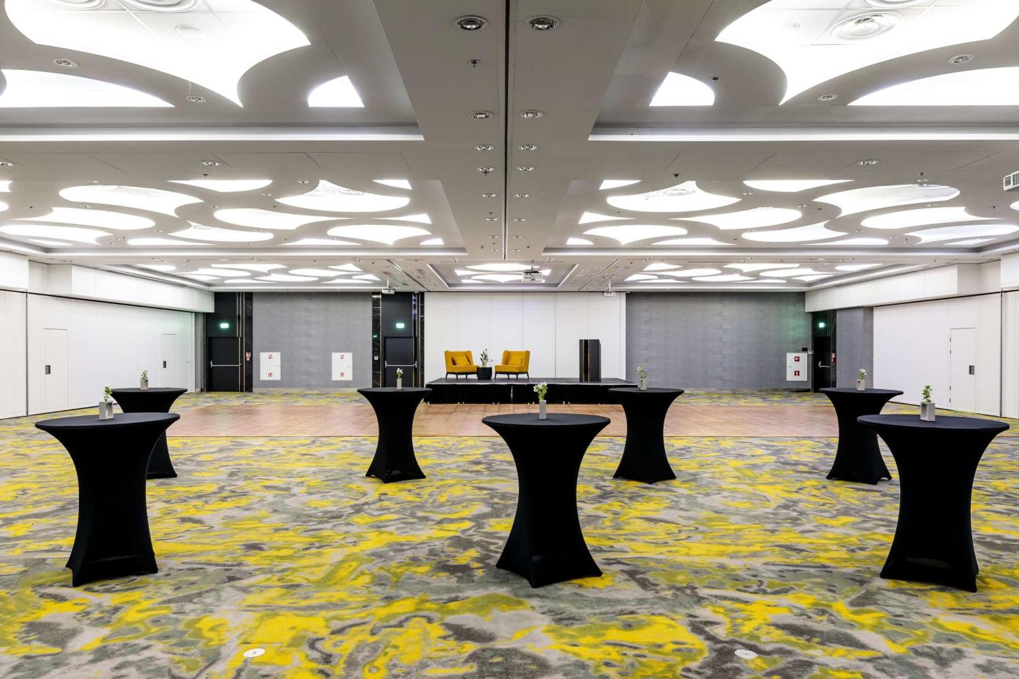 Radisson Blu Resort Świnoujście Екстериор снимка The photo shows a spacious interior room set up for an event. The flooring features a patterned design in gray with yellow accents. There are several black tall tables scattered throughout the area, and in the background, there are two large yellow a