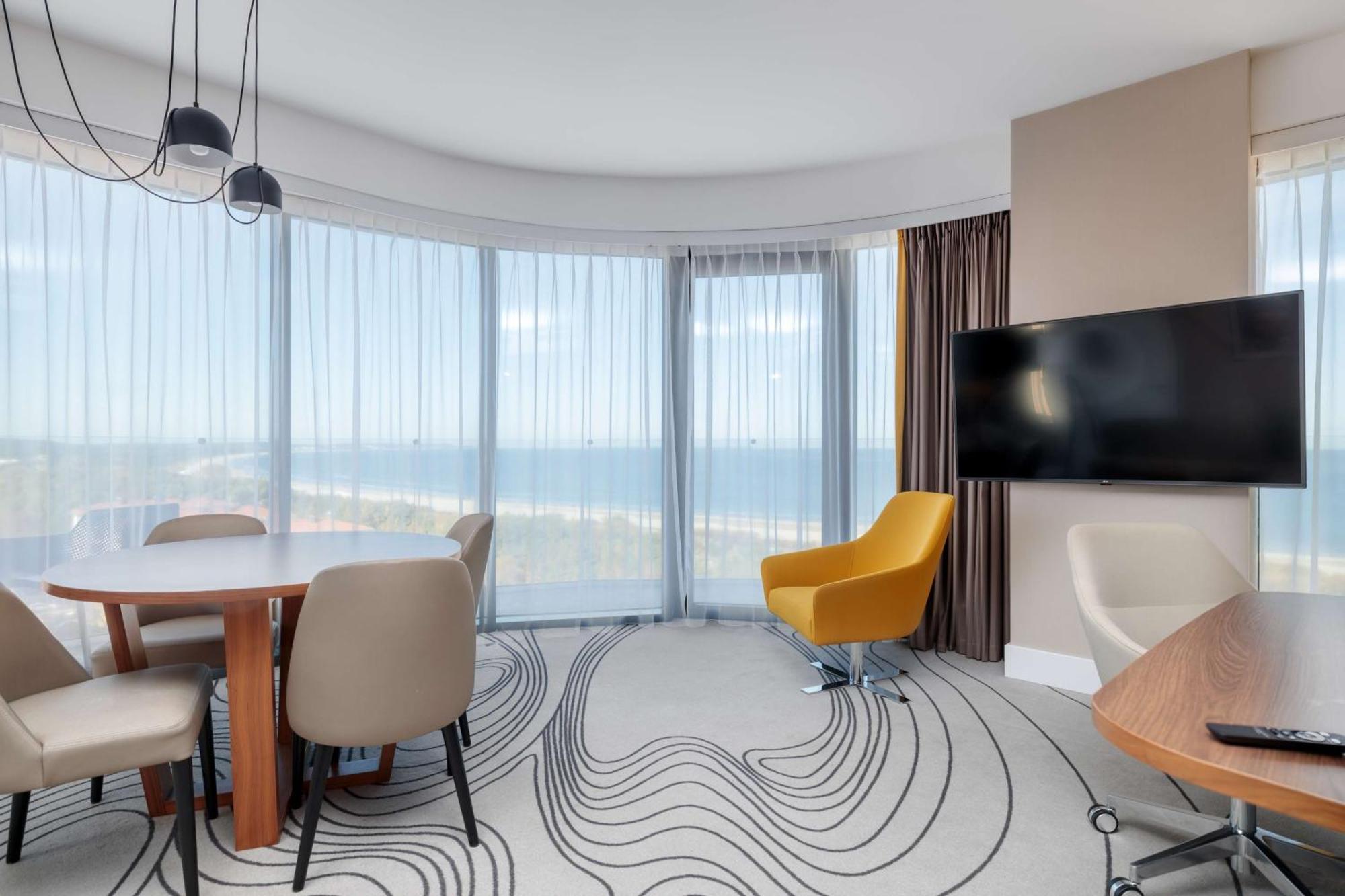 Radisson Blu Resort Świnoujście Екстериор снимка The photo shows a modern, stylish interior of a room with large windows that provide a panoramic view. The space features a round dining table surrounded by chairs, including a distinct yellow armchair. There is a flat-screen television mounted on th