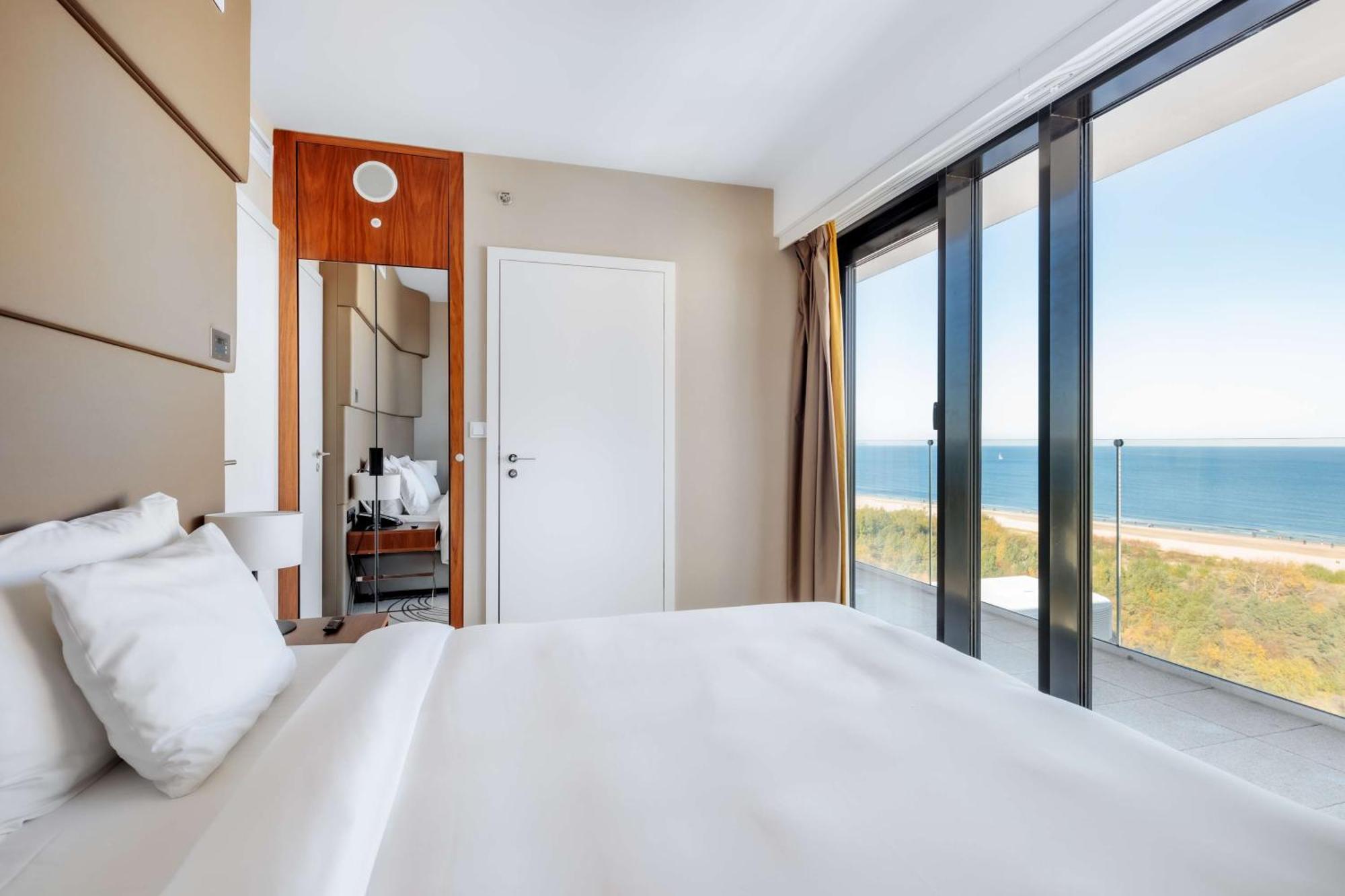Radisson Blu Resort Świnoujście Екстериор снимка The photo shows a modern hotel room with a bright and airy atmosphere. There is a large bed with white bedding positioned near a glass wall that offers a view of the ocean. The room features neutral-colored walls and wooden accents, enhancing its con
