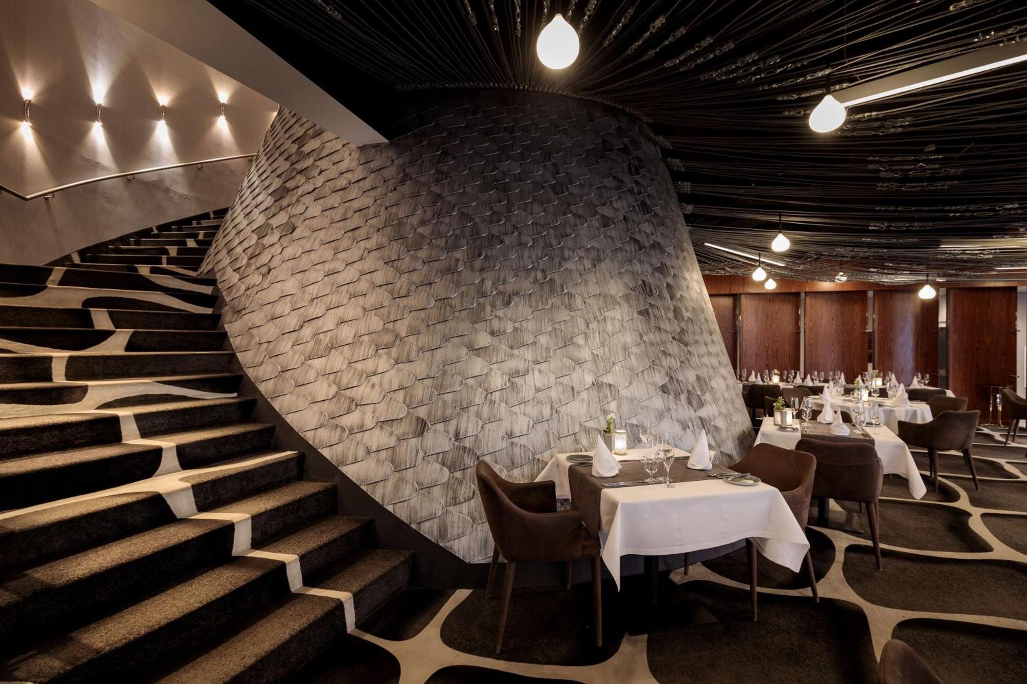 Radisson Blu Resort Świnoujście Екстериор снимка The photo shows the interior of a stylish restaurant. In the foreground, there are elegantly set tables with white tablecloths and dark chairs. A unique, curving stone wall adds a distinct architectural feature, and a grand staircase with a textured 
