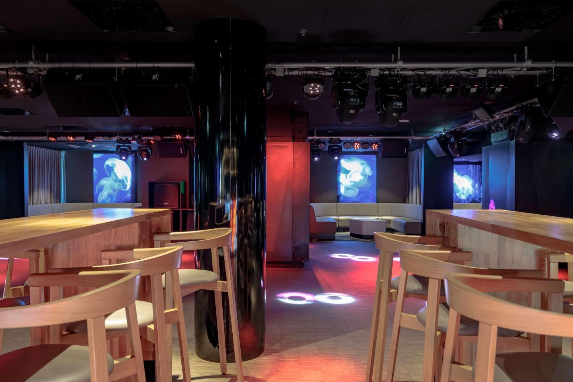 Radisson Blu Resort Świnoujście Екстериор снимка The photo shows an interior of a modern venue, likely a bar or club. The space features a central black column and light-colored wooden bar stools arranged around a bar area. On the walls, there are large screens displaying vibrant visuals, possibly 