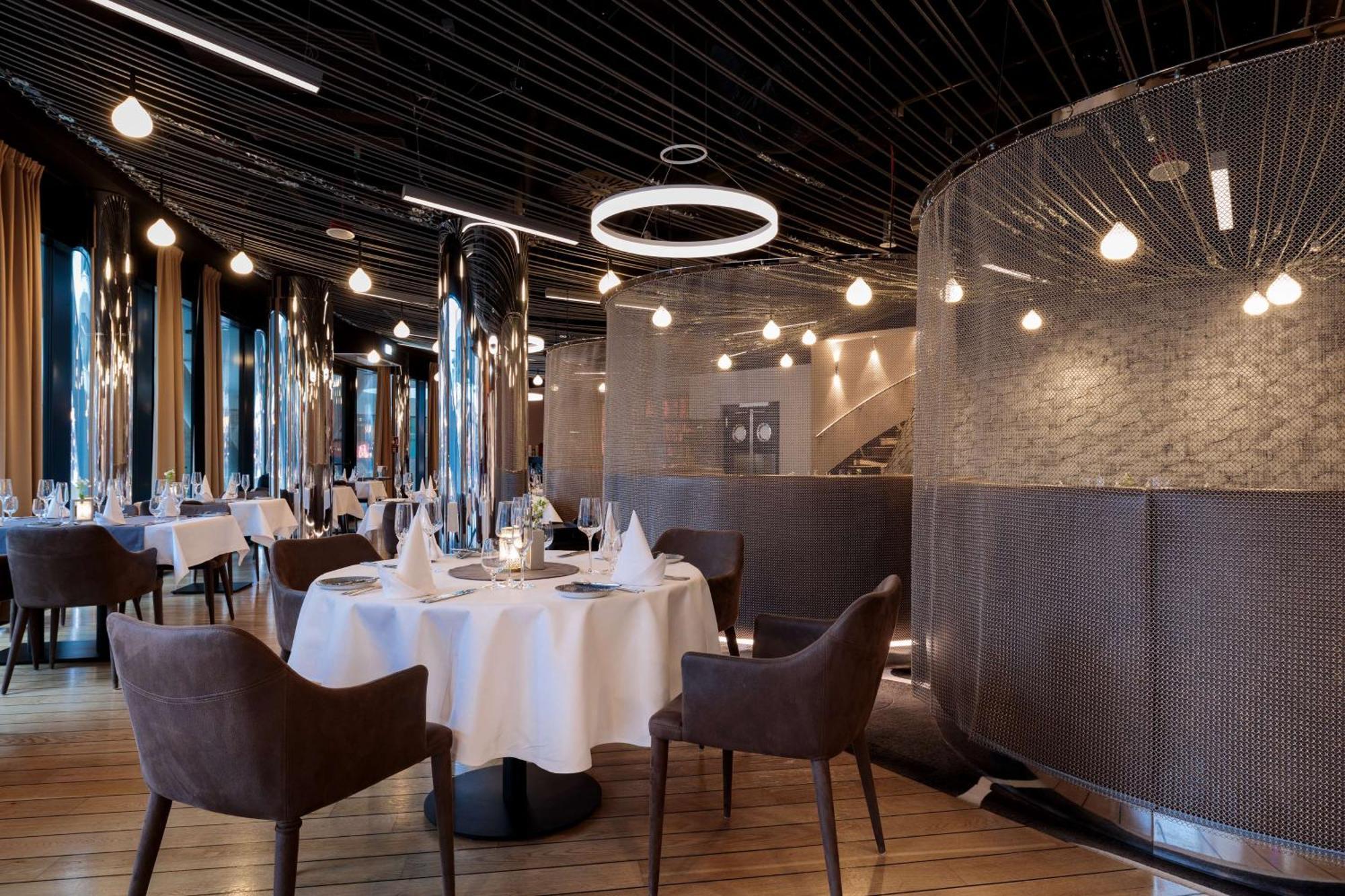 Radisson Blu Resort Świnoujście Екстериор снимка The image shows a modern restaurant interior that features a stylish design. There are several round tables with white tablecloths. The seating consists of brown, upholstered chairs. The space is illuminated by various hanging lights, including a dis